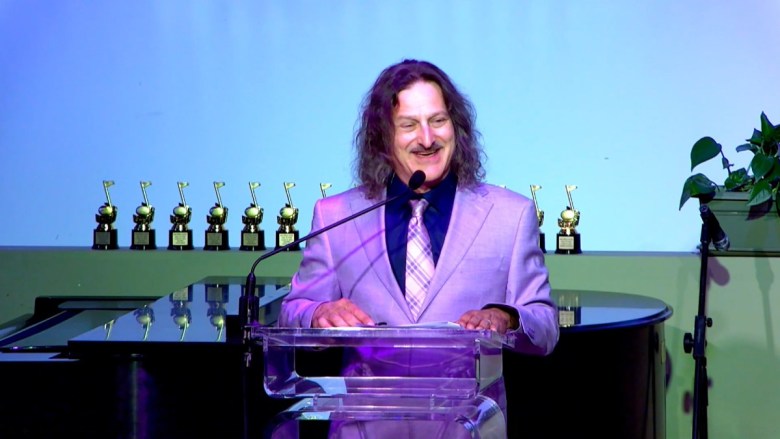 A person in a purple suit speaks at a podium with trophies and a piano behind them.
