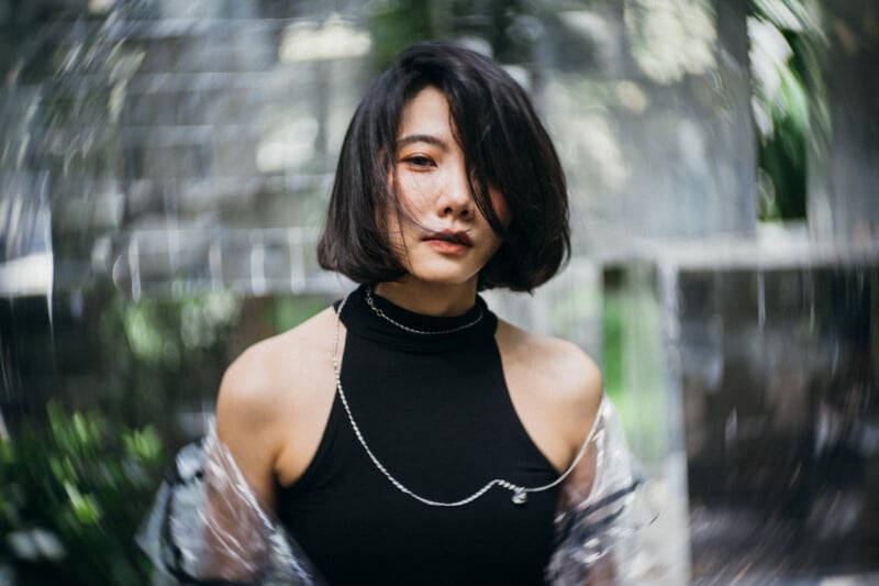 A woman with short dark hair in a black sleeveless top stands outdoors. The background is blurred in a circular pattern, drawing focus to her calm expression.