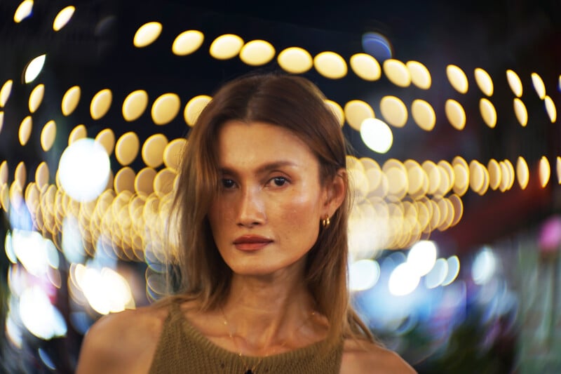 A woman with light brown hair and a neutral expression stands in front of blurred, circular string lights at night, creating a bokeh effect in the background. She wears a sleeveless top and gold hoop earrings.