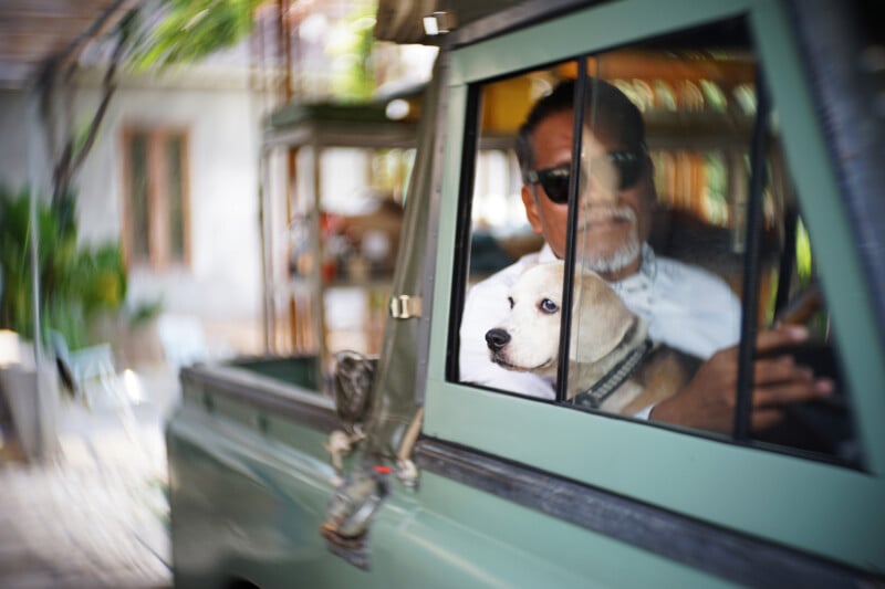 A man wearing sunglasses sits inside a green vehicle, looking out the window, with a white dog beside him also gazing outside. The background is blurred, suggesting a shallow depth of field.