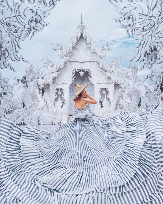 A woman in a wide, striped dress and sunhat stands with her back to the camera, facing an ornate, white temple with intricate carvings and details under a bright, blue sky.