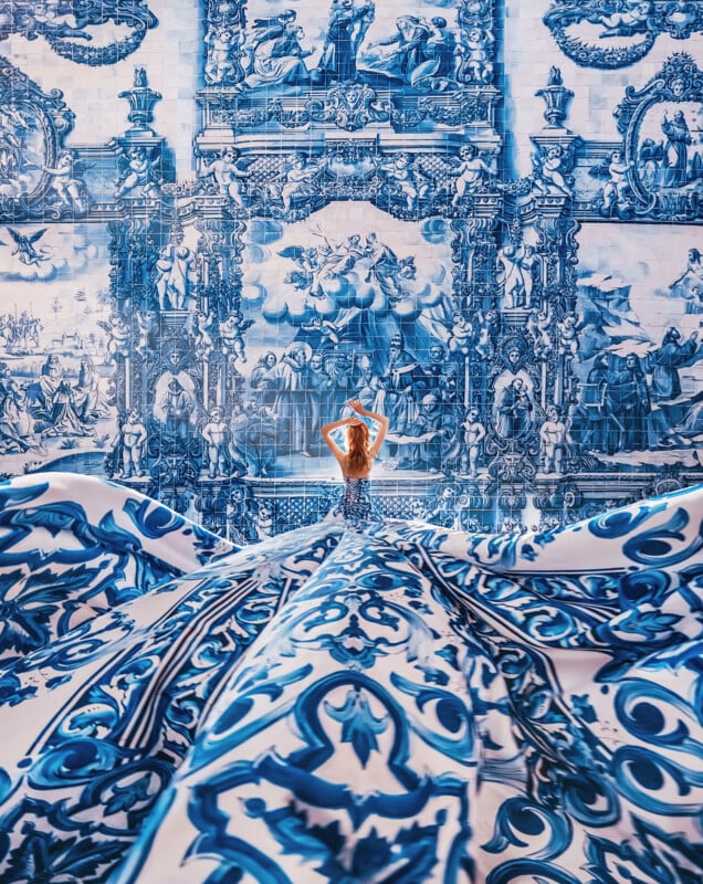 A woman in a flowing blue and white patterned dress stands with her arms raised, blending seamlessly into an ornate, blue-toned wall mural depicting classical scenes.