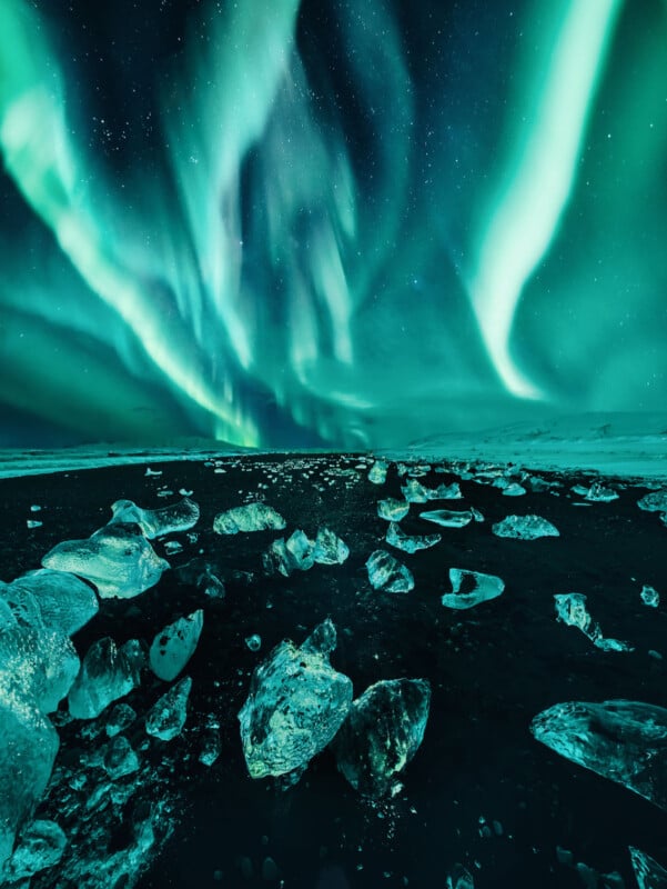 Shimmering green aurora borealis lights streak across a starry night sky above a dark, icy landscape scattered with large, translucent ice chunks.