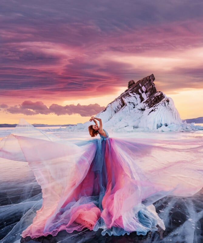 A woman in a flowing, pastel pink and blue gown poses gracefully on a frozen lake with ice formations, under a dramatic, colorful sunset sky with shades of purple, pink, and orange.