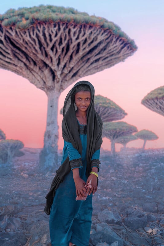 A young girl in a teal dress and dark shawl stands on rocky ground, smiling, with dragon blood trees and a pink sky in the background. She holds something in her hands and wears bracelets on her wrist.