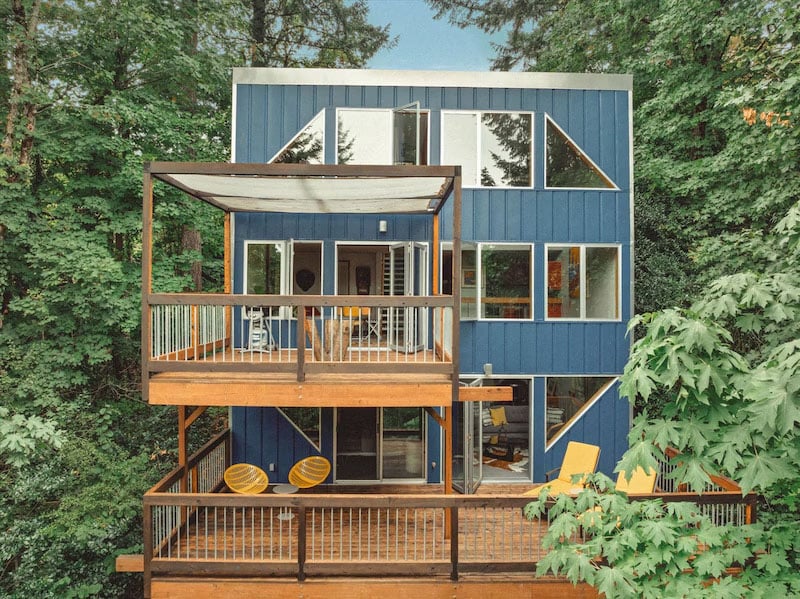 A modern blue house with large windows and two wooden balconies, surrounded by lush green trees. The balconies have seating areas, including yellow chairs, and the house has a geometric, contemporary design.