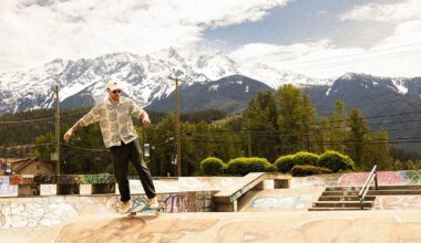A person skateboards on the edge of a graffiti-covered skatepark ramp, with lush trees and snow-capped mountains in the background under a partly cloudy sky.