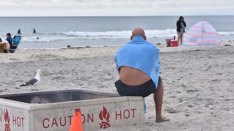A man warms himself at a fire pit after being in the ocean on the overcast day. Photo by Chris Stone