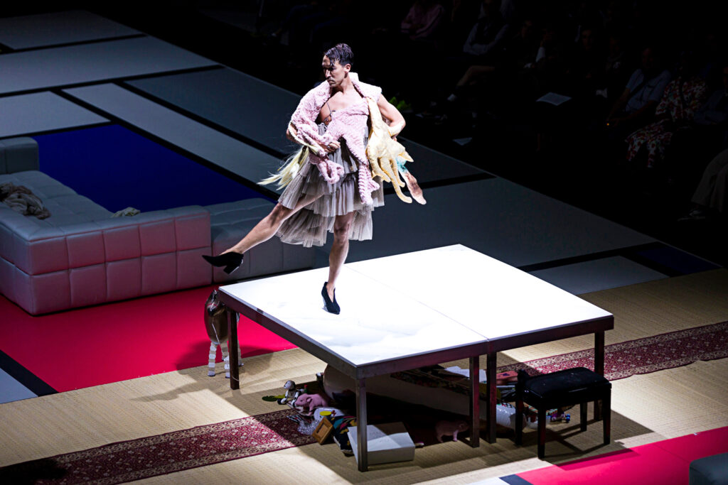 Atop a spotlit, minimalist table, a performer in heels, a short dress of layered tulle, and multiple knit garments layered over their shoulders poses with one foot extended side at forty-five. They look over their shoulder toward their extended foot, hands drawing back to their hips.