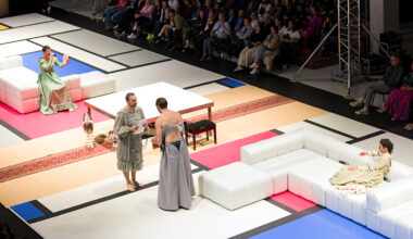 Two performers in old-fashioned evening gowns stand facing each other while holding sheets of paper on a long, rectangular Mondrian grid carpet. To either side of them are white sectional couches, with a performer in another similar dress seated on each. One lounges, while the other perches as they speak into a microphone while looking at a sheet of paper.