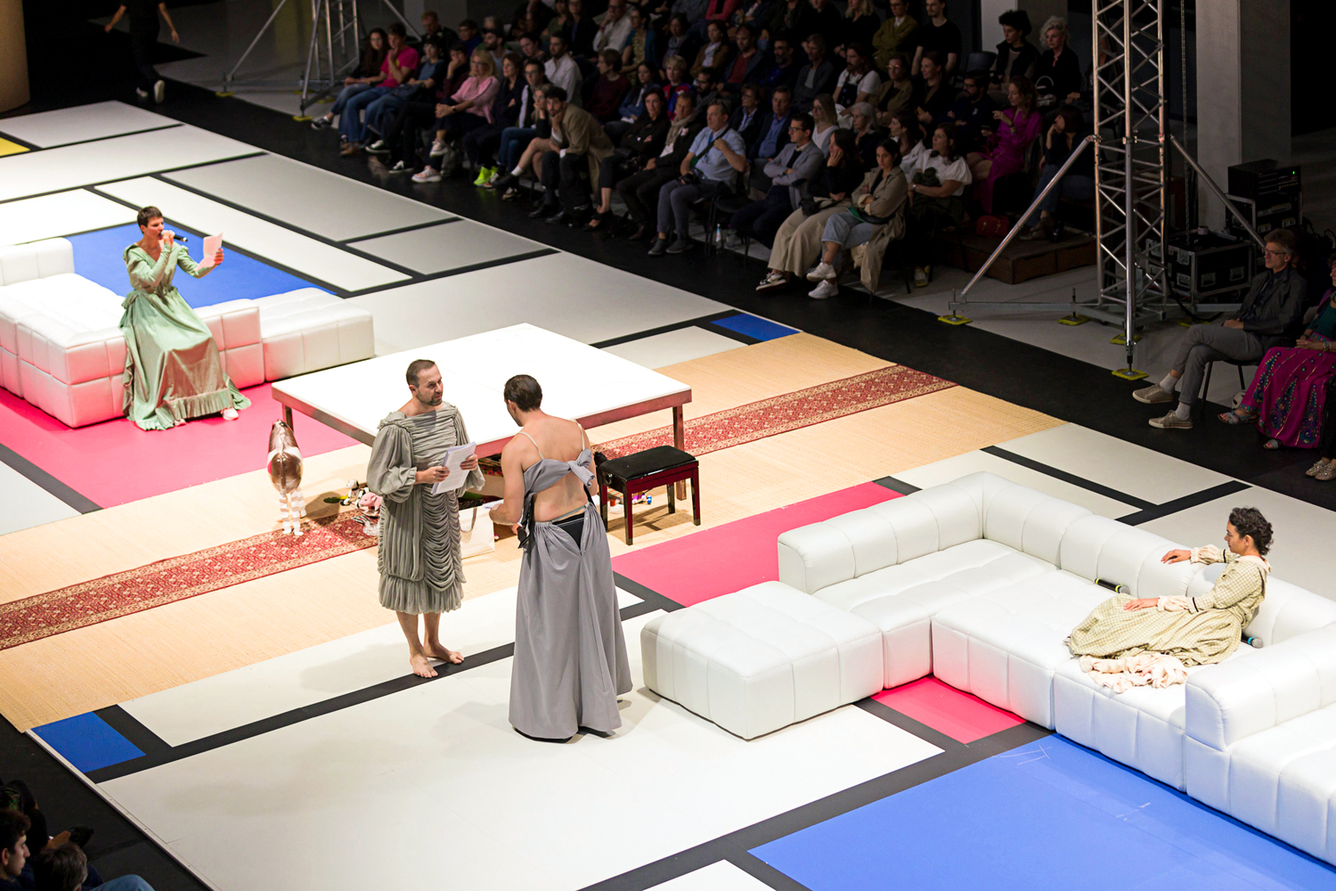Two performers in old-fashioned evening gowns stand facing each other while holding sheets of paper on a long, rectangular Mondrian grid carpet. To either side of them are white sectional couches, with a performer in another similar dress seated on each. One lounges, while the other perches as they speak into a microphone while looking at a sheet of paper.