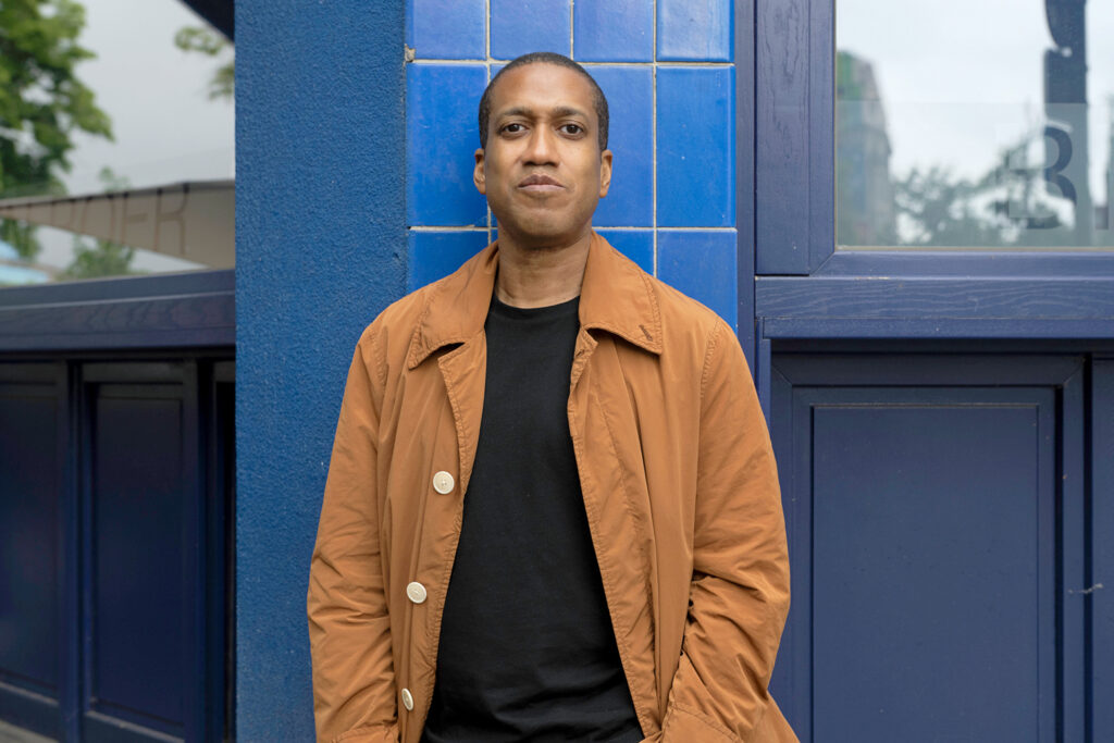 A portrait of Trajal Harrell. He looks seriously at the camera, hands in his pockets, while standing in front of a patch of blue tiles on a blue-painted wall. He wears an orange-brown jacket open over a plain black shirt.
