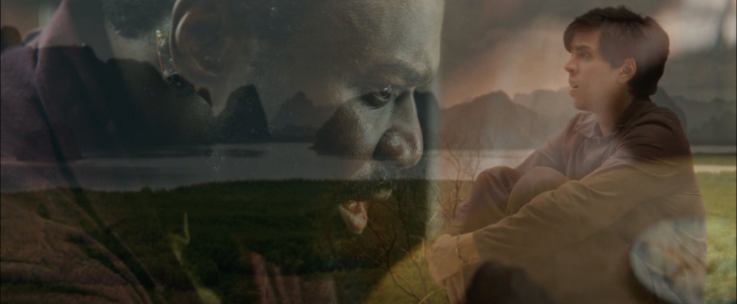A shot of a young man sitting in an open field next to a cliff. The shot is overlayed with the image of another man to illustrate that the two are communicating telepathically despite being located in different places.