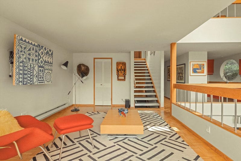 A modern living room with red lounge chairs, a patterned rug, a wooden coffee table, abstract art on the walls, and a staircase leading to an upper floor. Natural light fills the open space.