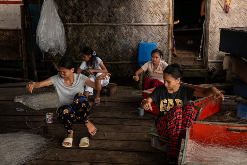Four women sit on a wooden floor inside a rustic building, wearing casual clothes, working with fishing nets. Woven bamboo walls, a red cooler, and plastic bags are visible in the background.