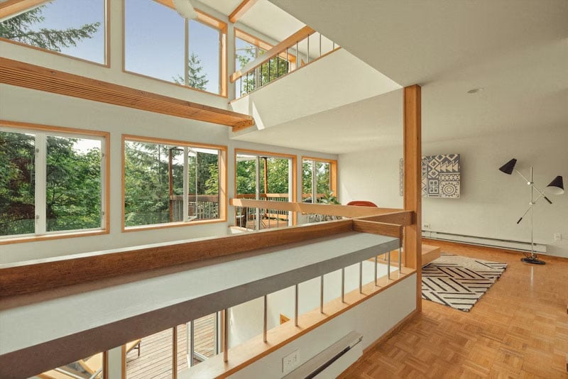 Modern, open-concept living space with large windows, wood accents, a patterned rug, and a floor lamp. Lush green trees are visible outside, filling the room with natural light.