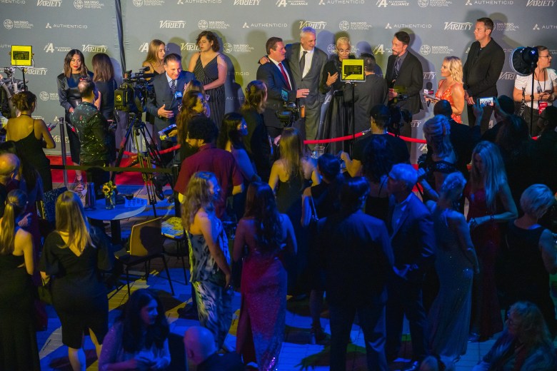 A crowd of people, gathered in clusters, is assembled on a red carpet for a film festival.