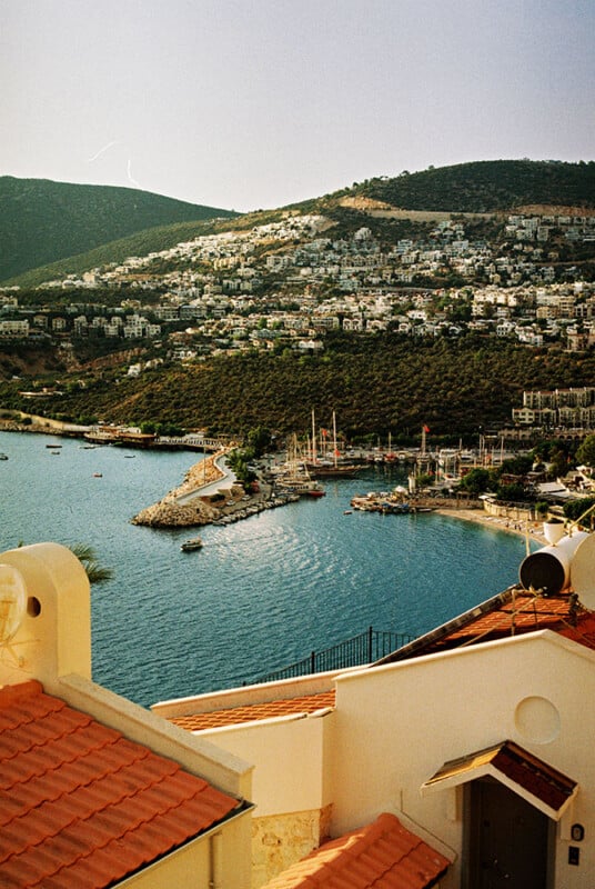 A coastal town with white buildings on green hillsides overlooks a calm bay with sailboats and a small marina. Terracotta rooftops appear in the foreground under a clear sky.