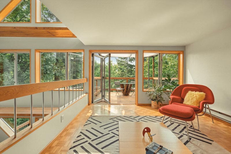 Bright living room with large windows, a modern orange chair with a yellow pillow, geometric rug, houseplant, and glass doors opening to a wooden balcony with trees outside.