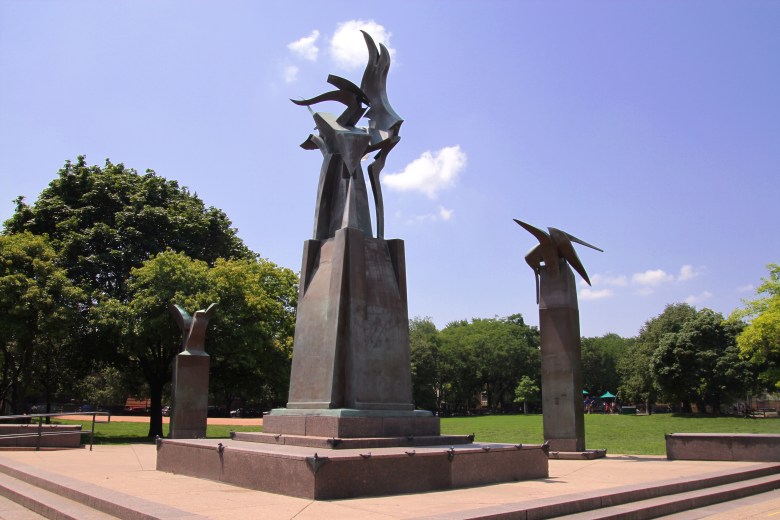 In a public park on a sunny day, a large monument by Richard Hunt is in the foreground. It features three metal columns with birdlike structures on top of three different sizes. The tallest, at the center of the image, is on a raised base. Trees are in the background behind a grass field.