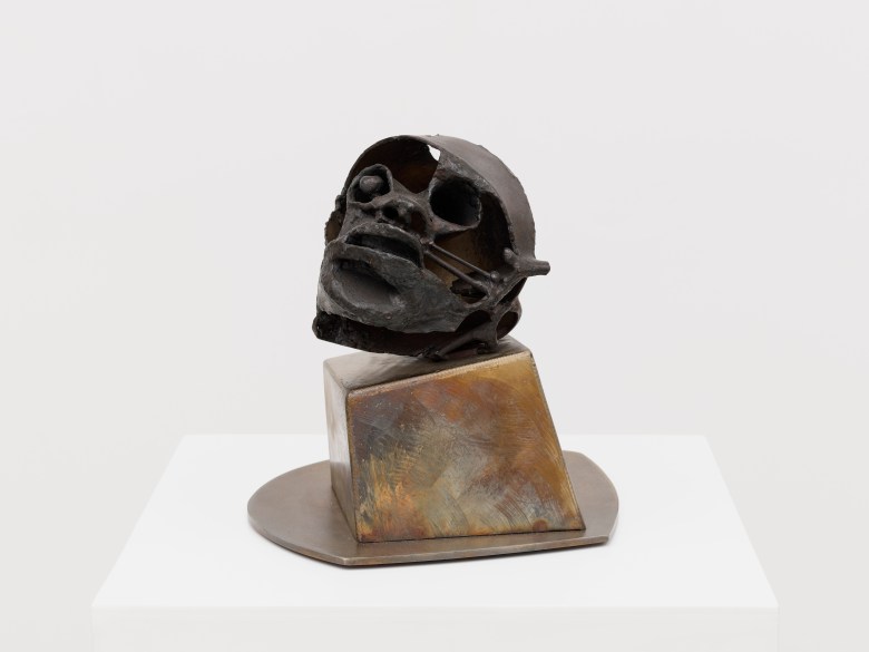 A small abstracted sculpture of a head sits on a metal, irregularly block-shaped base, on a white plinth. The head faces the camera. It has a helmet like band over its forehead and what appears to be a missing eye. The forehead and cheeks are also hollowed out.