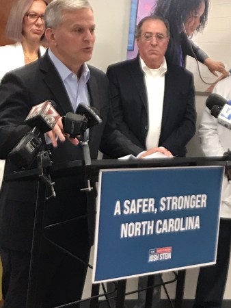 Gov. Josh Stein stands at a podium in Alliance Medical Ministry talking to reporters about pending Medicaid rate cuts. He is flanked by Pete Tannenbaum, executive director of Alliance Medical Ministry, and Jenna Beckham, an ob/gyn from Wake County.