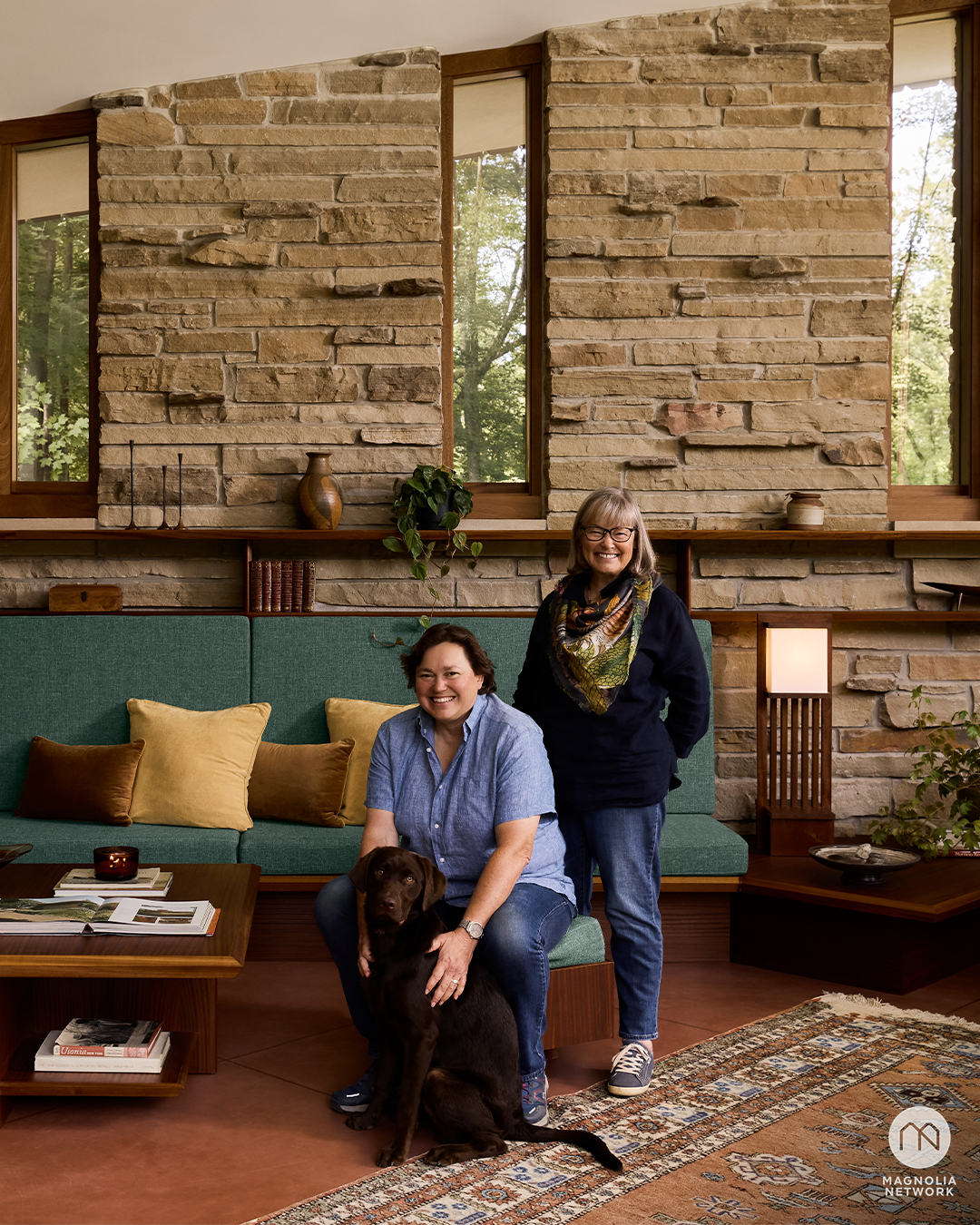 Two women pose in a mid-century modern living room with stone walls and large windows; one is seated with a dog, the other stands behind her.