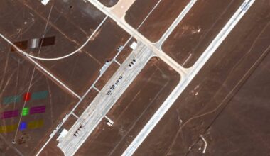 Satellite image of a desert airport runway and taxiway with aircraft lined up. Colorful rectangular markings are visible on the ground to the left of the runway complex.