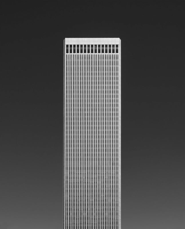 A tall, rectangular skyscraper with vertical white lines stands against a dark, gradient sky. The building's facade appears minimalist and symmetrical, and the image has a stark, modern feel.