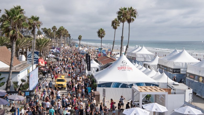 A crowded beachside event with people, palm trees, and white tents labeled "Nissan Super Girl Surf Pro."
