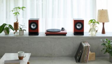 A minimalist setup featuring a red turntable flanked by two matching red speakers on a concrete ledge. The ledge also holds several small potted plants, a glass pitcher with water and green leaves, and a wooden table lamp with a beige shade. Below the ledge, there is a stack of vinyl records leaning against the wall. In the foreground, a white table with a brown and white coffee mug and a closed book is visible. The background consists of sheer white curtains allowing soft natural light.