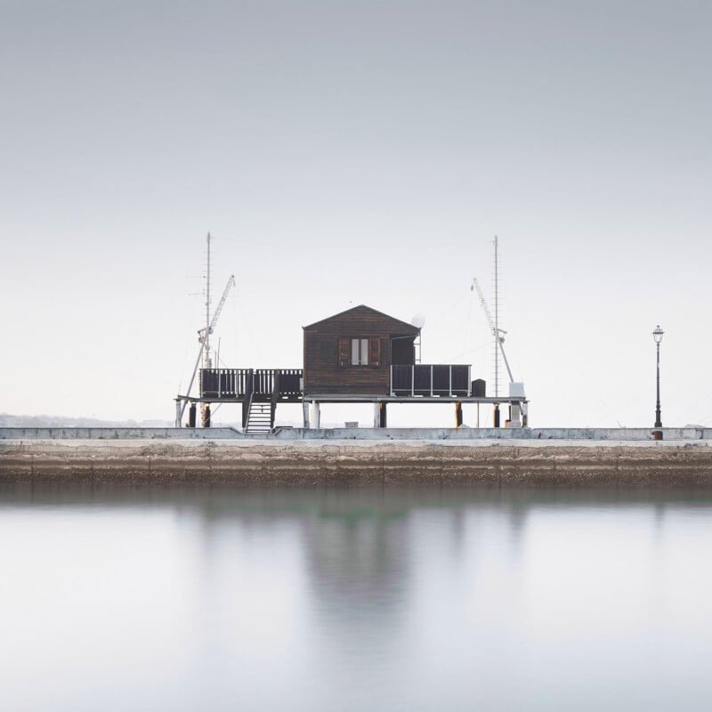 A small wooden house on stilts sits on a pier above calm water, flanked by fishing poles and a single streetlamp under a pale sky. The scene is minimalistic and serene.