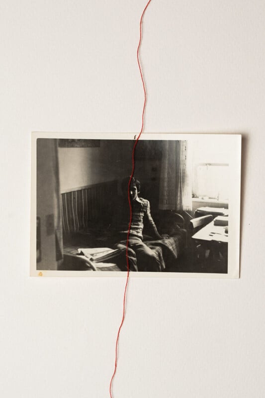 A black and white photo of a person sitting on a bed is pinned to a white wall. A red thread runs vertically, dividing both the image and the wall behind it in half.
