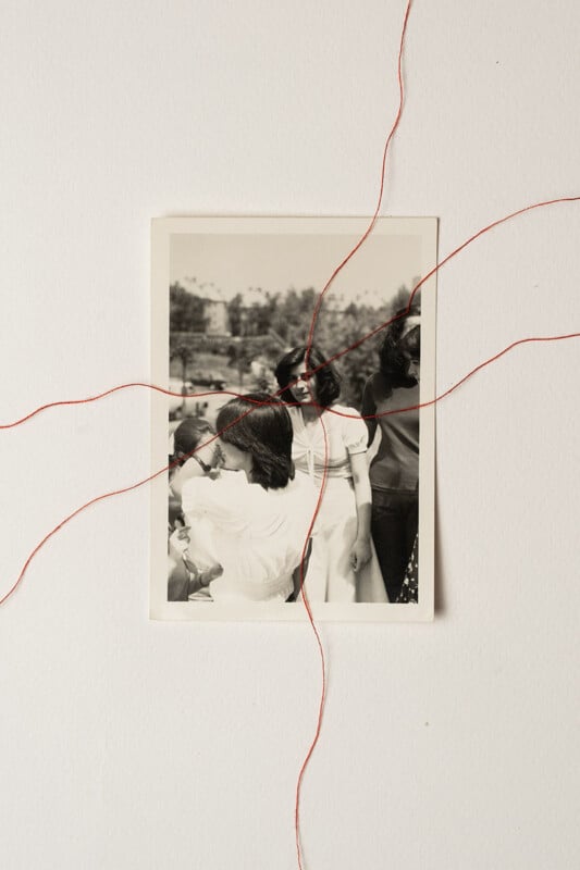 A black-and-white photo of three women is pinned to a white wall. Red threads are stitched through and around the photo, radiating outward, some passing across faces and joining at points on the image.