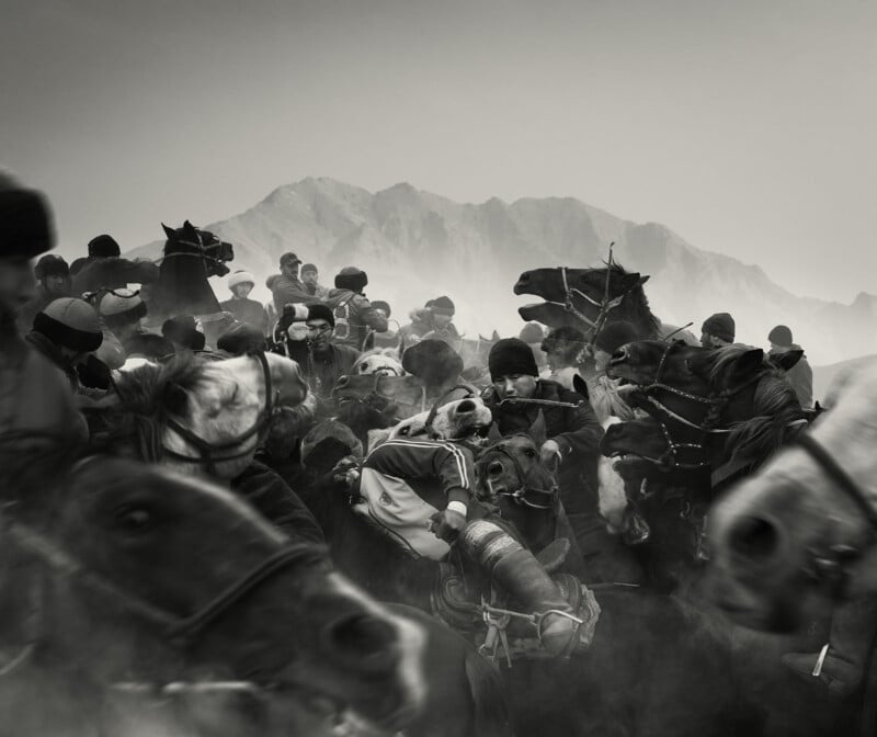 A group of men on horseback compete in a chaotic, dust-filled game, surrounded by mountains in the background; the scene is intense with tightly packed horses and riders.