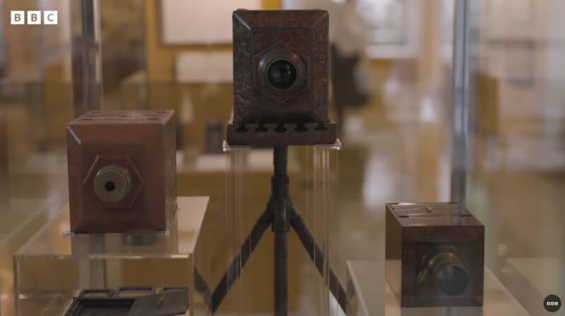 Three antique box cameras are displayed in a museum exhibit, each encased in glass. The camera in the center is mounted on a tripod, while the other two are placed on the surface beside it. BBC logo is visible in the top left corner.