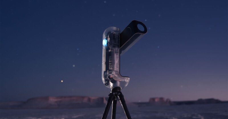 A modern, transparent telescope on a tripod stands outdoors at dusk, with a clear sky and distant mesas in the background. Stars are visible in the sky, and the scene has a calm, twilight atmosphere.