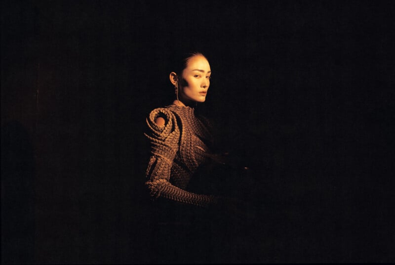 A woman in a textured, futuristic outfit stands in soft, dramatic lighting against a dark background, creating a moody and artistic portrait.