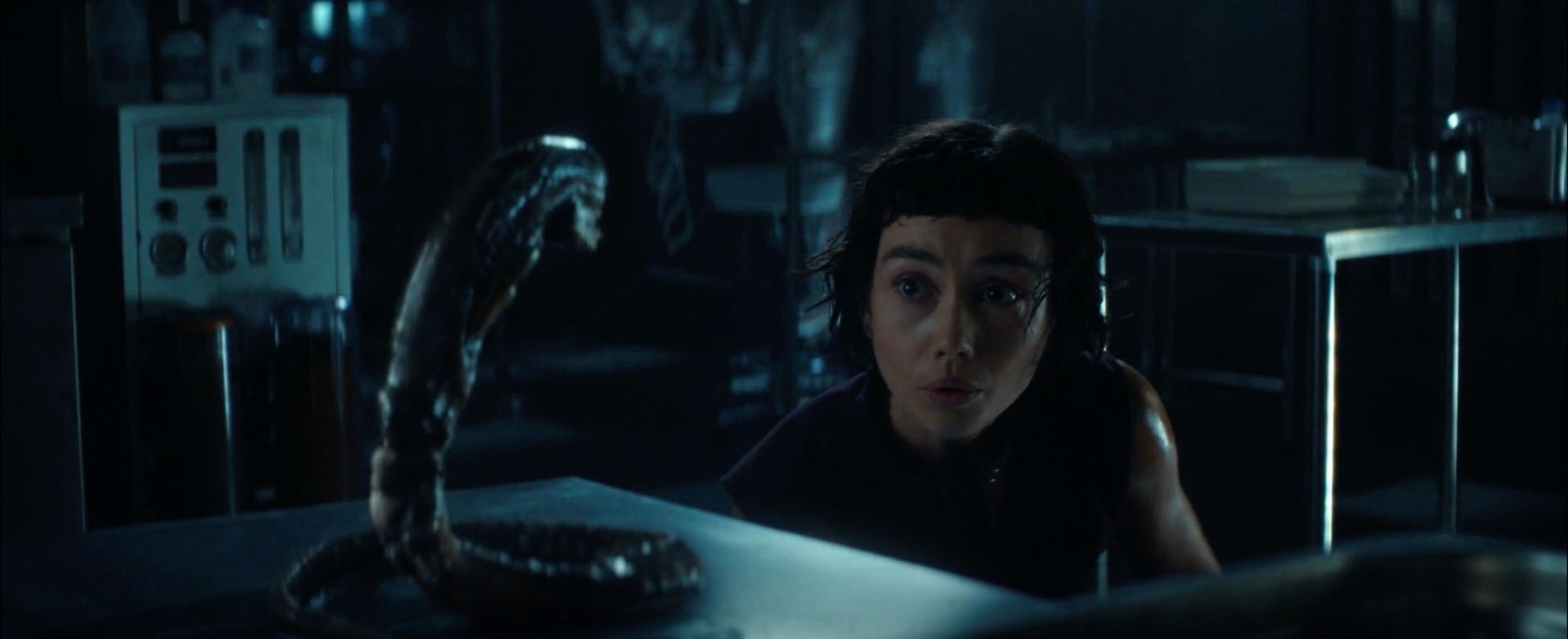 A woman crouching low to the ground to make cooing noises at a snake-like alien, which is setting on a metal examination table.