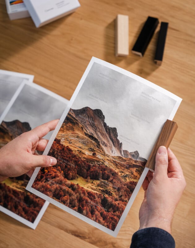 A person holds a landscape photo of mountains and autumn trees on a wooden table, using a wooden clip. Other prints and wood samples are also visible on the table.