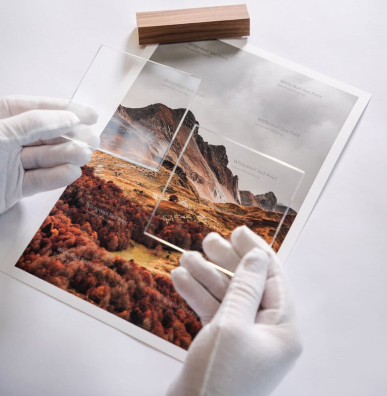 A person wearing white gloves holds clear acrylic sheets above a printed landscape photo of mountains and autumnal trees, demonstrating how the sheets overlay the image. A rectangular wooden block rests above the print.
