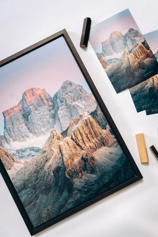 A framed photograph and prints of rugged, sunlit mountain peaks on a white surface, with wooden frame corners and tools placed nearby.