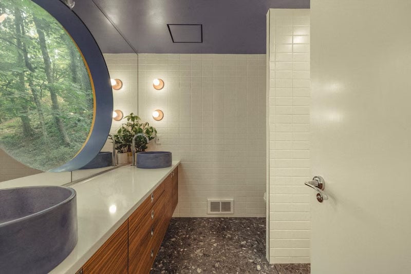 A modern bathroom with a blue ceiling, large round mirror reflecting trees, white tiled walls, wooden vanity with drawers, two blue vessel sinks, round wall lights, and a potted plant on the counter.