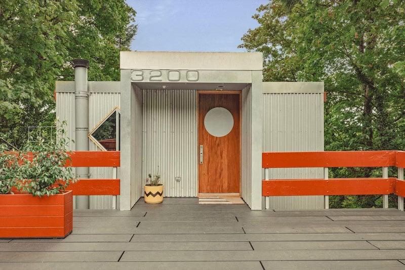 A modern house entrance with a red wooden door, vertical siding, and the number 3200 above. Red horizontal railings, potted plants, and trees in the background complete the scene.