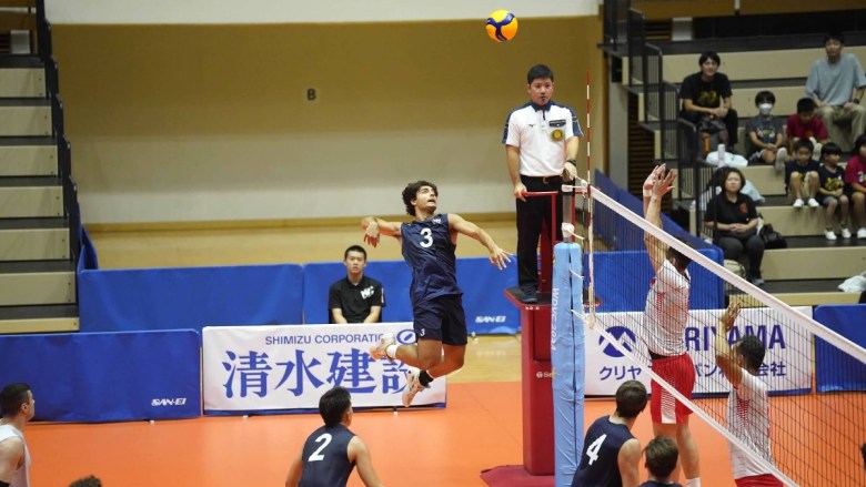 A volleyball player in a blue jersey spikes the ball, while two players in white and red jerseys attempt to block it, with an official observing the play.