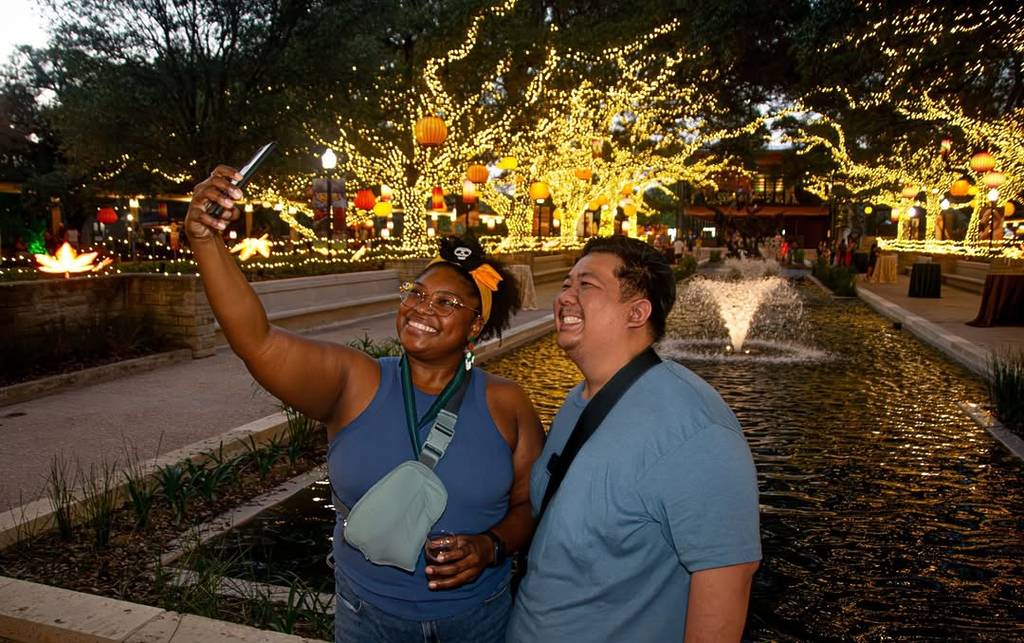 Image shows a couple taking a photo of themselves with a lit up tree in the background at the Houston Zoo