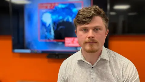 A man in a white shirt sits in an office, in front of a large monitor that has CCTV footage of a sewerage pipe