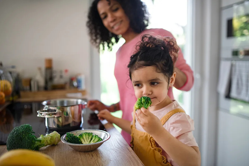 Happy multiracial girl preparing broccoli for lunch with her mother in kitchen.