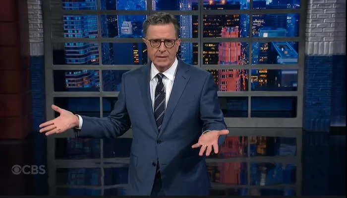 A person in a suit stands with arms open on a talk show set, cityscape visible through the window behind them