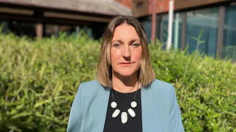 A woman with brown hair, green jacket and clack shirt stands in from of an office building.